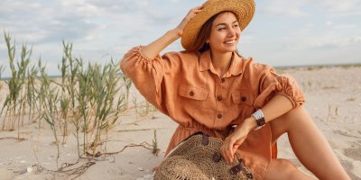 Žena ve volných košilových šatech a klobouku sedí na písečné pláži