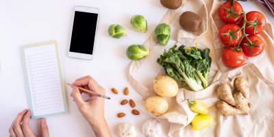 Sepisování nákupního seznamu vedle telefonu a látkové tašky s rozličnou zeleninou