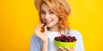 Žena v klobouku drží misku s třešněmi a jednu třešeň se chystá sníst