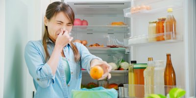 Žena stojí u otevřené lednice, mračí se, zacpává si nos a vyhazuje mandarinku do koše