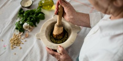 Starší žena v hmoždíři připravuje pesto, před ní jsou piniové ořechy, česnek, bazalka, olivový olej a parmazán