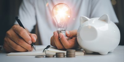 Muž si zapisuje poznámky do sešitu, v ruce drží rozsvícenou žárovku, před ním jsou vyskládané mince a kasička ve tvaru prasátka.