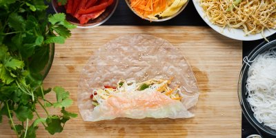 Balení letního závitku do rýžového papíru na dřevěném prkénku, misky s ingrediencemi (nudle, zelenina) v okolí