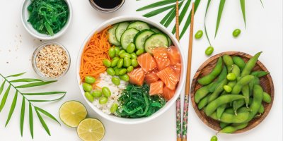 Poke bowl s lososem, řasami, rýží, edamame boby, mrkví a okurkou, vedle je limetka, lusky edamame, sezam, hůlky