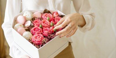 Žena drží dárkový box, ve kterém jsou květiny, makronky a kosmetika