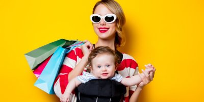 Žena s brýlemi má v na sobě dítě v nosítku a drží nákupní tašky