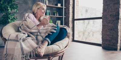 Žena zabalená do deky, sedící v křesle s hrnkem čaje