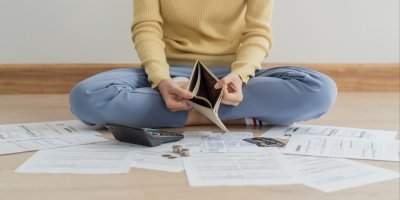 Žena sedí na zemi, drží prázdnou peněženku a kolem ní jsou faktury, kalkulačka, platební karta a mince