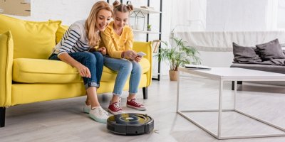 Žena s dcerou sedí na gauči a koukají na robotický vysavač