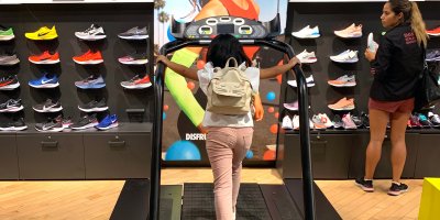 Žena na běžícím páse v obchodě před stěnou, na níž jsou vystavené různé tenisky