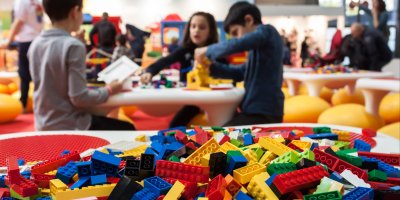 Hromada kostiček LEGO, v pozadí děti skládající stavebnici v obchodě