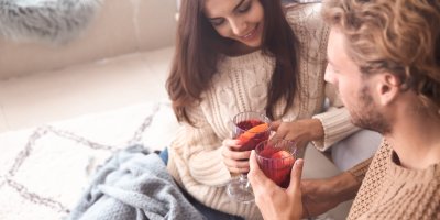 Muž a žena ve svetrech sedí doma na zemi pod dekou a v ruce drží svařák
