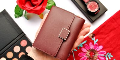 Žena drží malou hnědou peněženku v dlani, v pozadí je paletka očních stínů, hodinky, květina a barevné oblečení