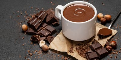 Hustá horká čokoláda v hrnku, kolem jsou kostičky čokolády a lískové ořechy