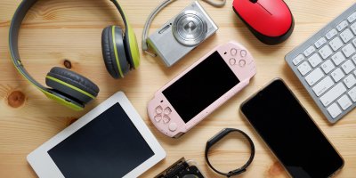 Různé druhy elektroniky ležící na stole – sluchátka, telefon, fotoaparát, myš, klávesnice, tablet a herní konzole