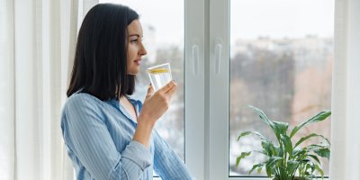 Žena stojí u okna a v ruce drží sklenici s vodou a plátkem citronu