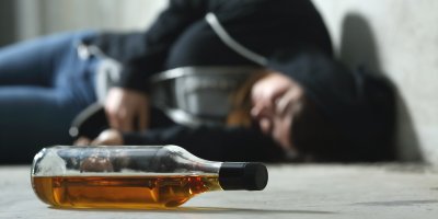 Detail ležící lahve s alkoholem, v pozadí muž v kapuci ležící na zemi