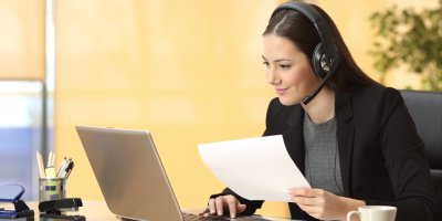 Žena má na hlavě sluchátka s mikrofonem, v ruce drží papír a dívá se do notebooku