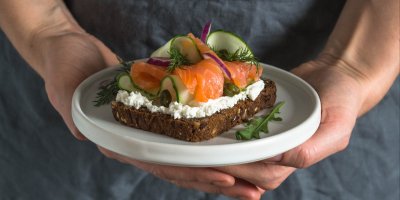 Člověk drží talíř s jedním dánským chlebíčkem s lososem, okurkou, cibulí a koprem