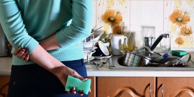 Žena stojí s houbičkou v ruce u špinavého nádobí v dřezu