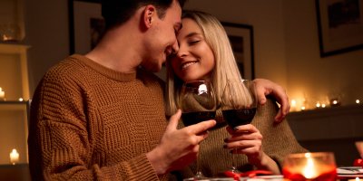 Muž a žena sedí u romanticky prostřeného stolu, opírají se o sebe a v ruce drží sklenice s červeným vínem