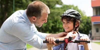 Muž dává dítěti na koloběžce přilbu
