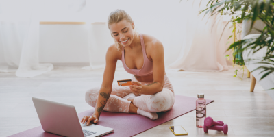 Žena sedí ve sportovním oblečení na fitness podložce a objednává něco na notebooku