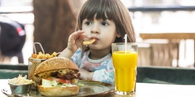 Holčička sedí u stolu s hamburgerem, hranolky a džusem