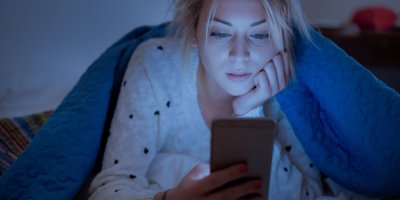 Žena leží na břiše přikrytá peřinou a dívá se do mobilu