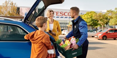 Muž dává do auta Tesco bedýnku s potravinami, sleduje jej žena s chlapcem