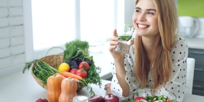 Žena pije vodu s okurkou, vedle sebe má košík se zeleninou a mísu se salátem