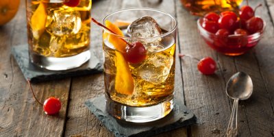 Dva koktejly Old Fashioned s ledem, pomerančem a třešněmi, v pozadí miska s koktejlovými třešněmi a lžička