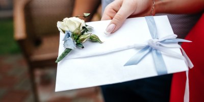 Ženská ruka drží obálku ovázanou modrou stužkou a ozdobenou květinou