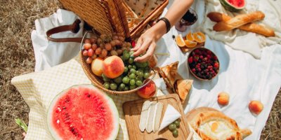 Otevřený koš na piknikové dece, mísa ovoce, půlka melounu, nakrájené sýry, sendviče, bagety