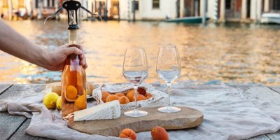 Deka na břehu řeky, člověk otevírá lahev vína, na prkénku jsou dvě sklenice, sýr a meruňky