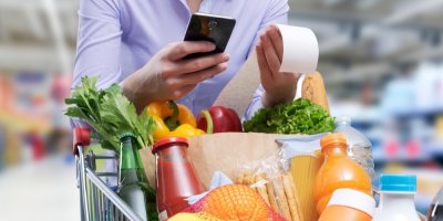 Člověk má plný vozík nakoupených potravin a prohlíží si účtenku a mobilní telefon