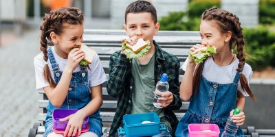 Dvě dívky a jeden chlapec sedí na lavičce a všichni jedí sendvič