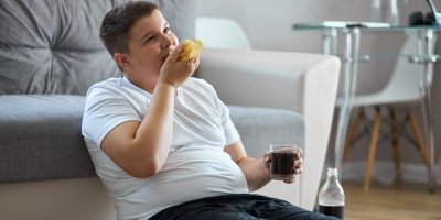 Obézní chlapec jí nezdravé jídlo a pije slazený nápoj u televize