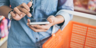 Žena v modré džínové košili a slunečních brýlích používá mobilní telefon k porovnání ceny a drží oranžový nákupní košík