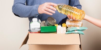 Muž drží krabici s trvanlivými potravinami, další člověk mu podává lahev oleje