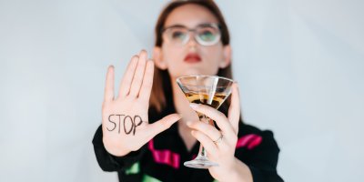 Žena drží sklenici alkoholu, druhou ruku má zdviženou a na dlani má nápis stop