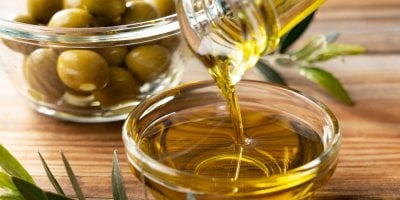 Nalévání olivového oleje do skleněné misky, v pozadí olivy