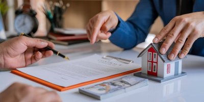 Pročítání smlouvy, týkající se koupě nemovitosti. Na stole vedle smlouvy je umístěný model domu a pár bankovek