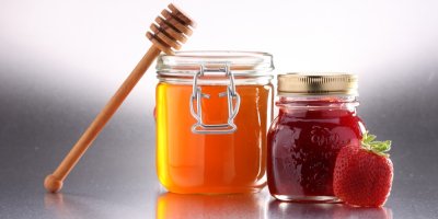 Sklenice medu a sklenice jahodové marmelády, vedle níž je jahoda