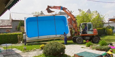 Bagr drží ve vzduchu bazén, pod ním stojí dva muži