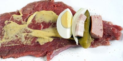 Plátek hovězího masa, potřený hořčicí, na plátku položené vejce, slanina, okurka a gothaj
