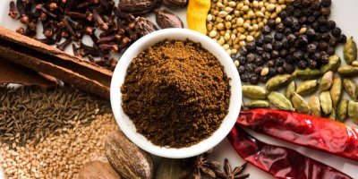 Koření garam masala v bílé misce, kolem ní koření, z něhož se vyrábí