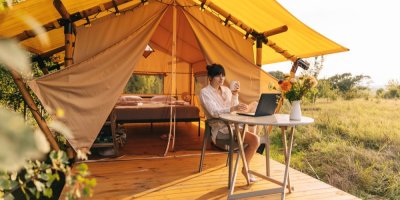 Žena sedí před glampingovým stanem na židličce, popíjí kávu a pracuje na laptopu.