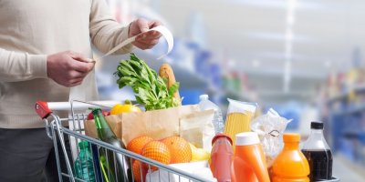 Muž drží v ruce lístek z nákupu a před ním je plný vozík potravin