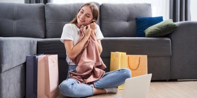 Mladá žena si přikládá k tváři nové oblečení, okolo jsou nákupní tašky